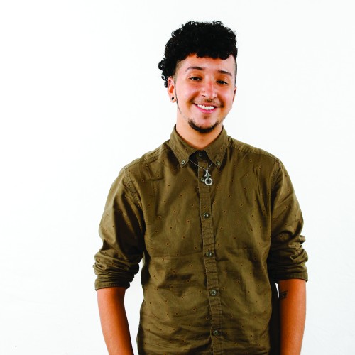 A person with an olive green button up is seen smiling at the camera, arms relaxed to the sides.